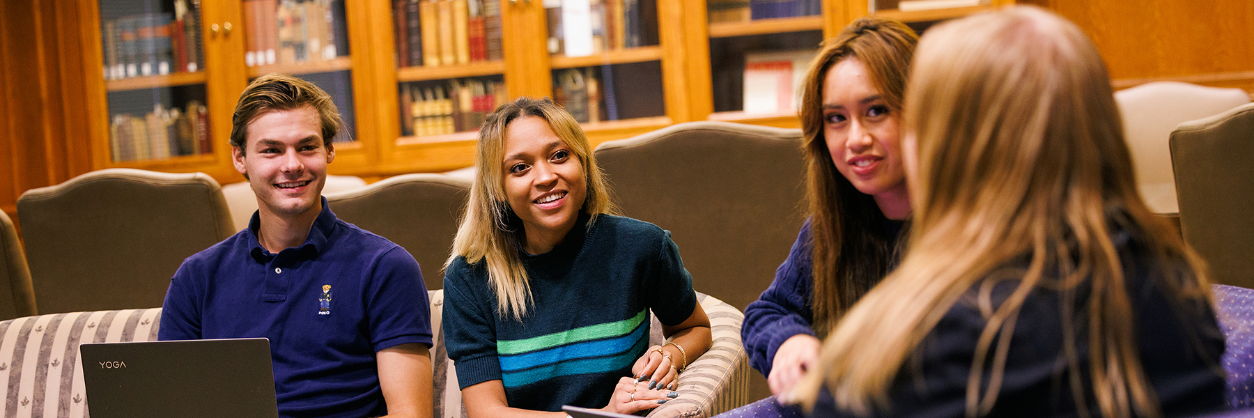 A diverse group of college students sit together in a library setting. They are engaged in conversation and smiling, appearing to be collaborating on a project or studying. A bookshelf is visible in the background.