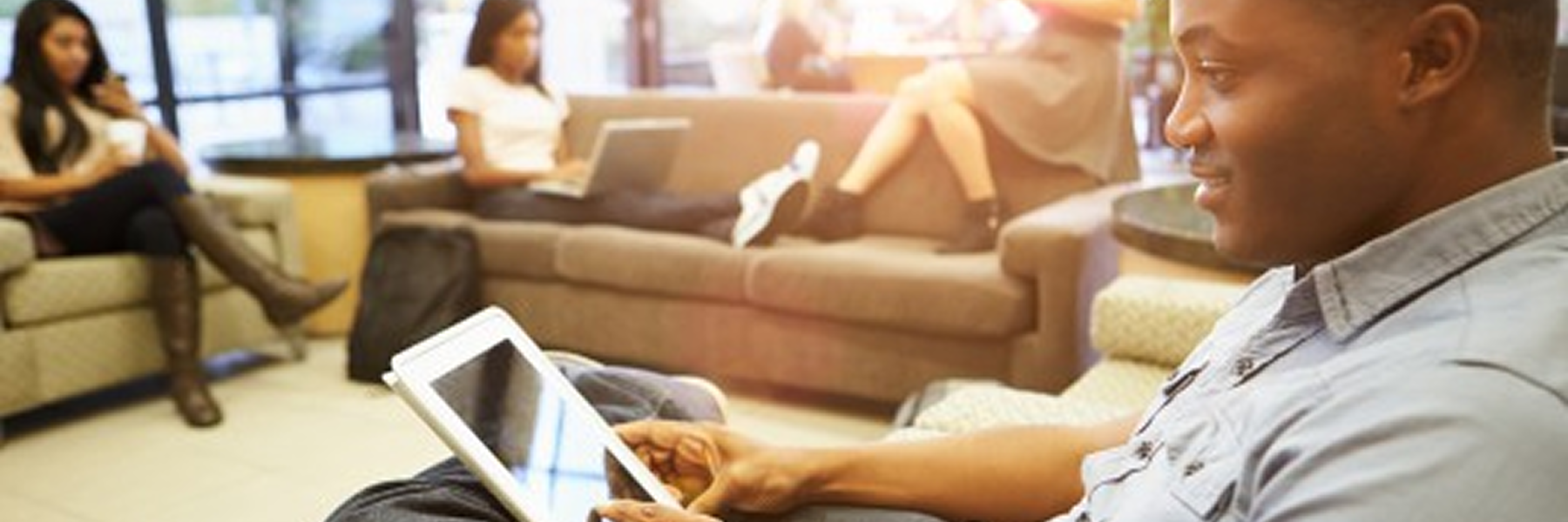 A side-angle photo of a young man, who is Black, sitting in a chair and looking at a tablet in his lap. He is smiling and appears to be in a comfortable lounge or study area with other students. In the background, two students are sitting on a couch and a third is sitting in a chair. The space is well-lit from a window.